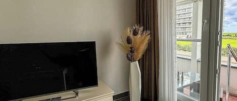 Uma sala de estar moderna com uma televisão de tela plana em um móvel, um vaso com plantas secas e uma porta de vidro deslizante que dá para uma varanda.