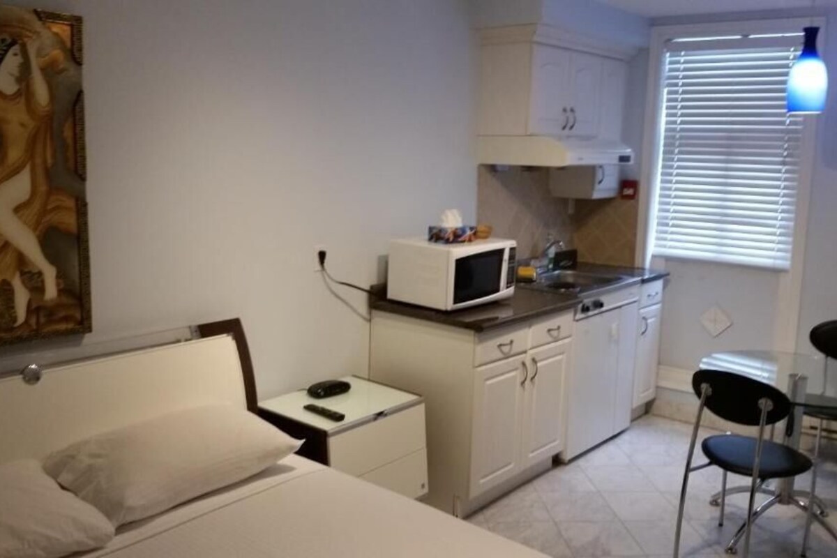 Een kleine kamer met een bed, een klein nachtkastje, een magnetron, een wastafel en een stoel.