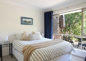 A bedroom with a bed, a bedside table with a lamp, a window with curtains, and a view of greenery outside. - Seaside Escape | Ocean Views & Spa Bath (Denhams Beach)