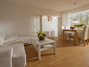 A modern living room with a sofa set, a dining table with chairs, and a coffee table with a flower arrangement. - Holiday Apartment in Norderney With Wi-fi (Norderney)