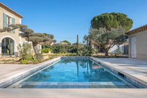 A swimming pool with a clear blue water surface, surrounded by a landscaped garden with various trees and shrubs, and adjacent to a building with a tiled roof. - Comfortable Villa in Saint-remy-de-provence (St.-Rémy-de-Provence)