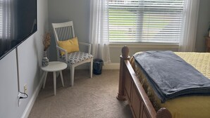 A bedroom with a bed, a chair, a television, a window with blinds, and a ceiling fan.