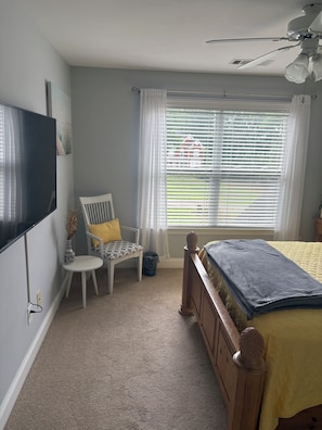 Un dormitorio con cama, una silla, televisor, ventana con persianas y ventilador de techo.