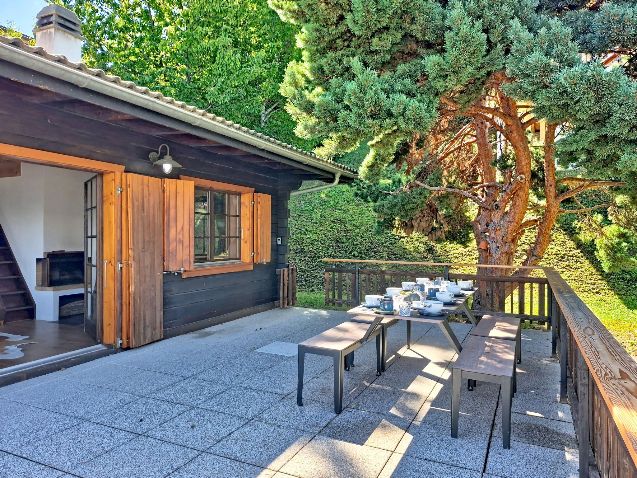 A modern outdoor patio with a dining table set for four, surrounded by a wooden deck and a large tree.