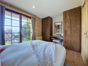 Ein Schlafzimmer mit einem großen Bett, einem Holzschrank, einem Schreibtisch mit Spiegel und Blick auf Bäume durch ein Fenster.