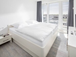 A modern hotel room with a large bed, a bedside table, a desk, and a view of the city through large windows. - Holiday Apartment in Norderney With Sauna (Norderney)