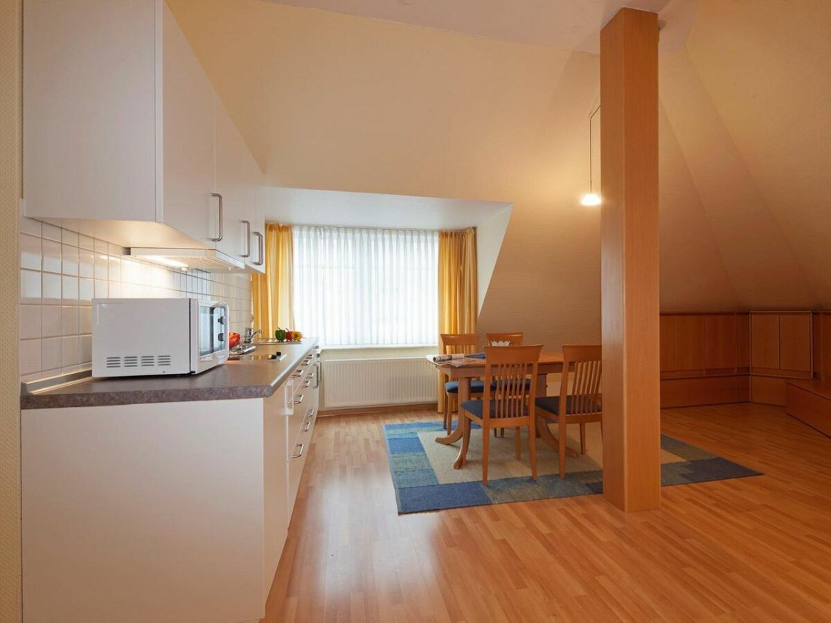 A compact kitchen with a microwave, stove, and sink, a dining area with a table and chairs, and a large window with curtains.