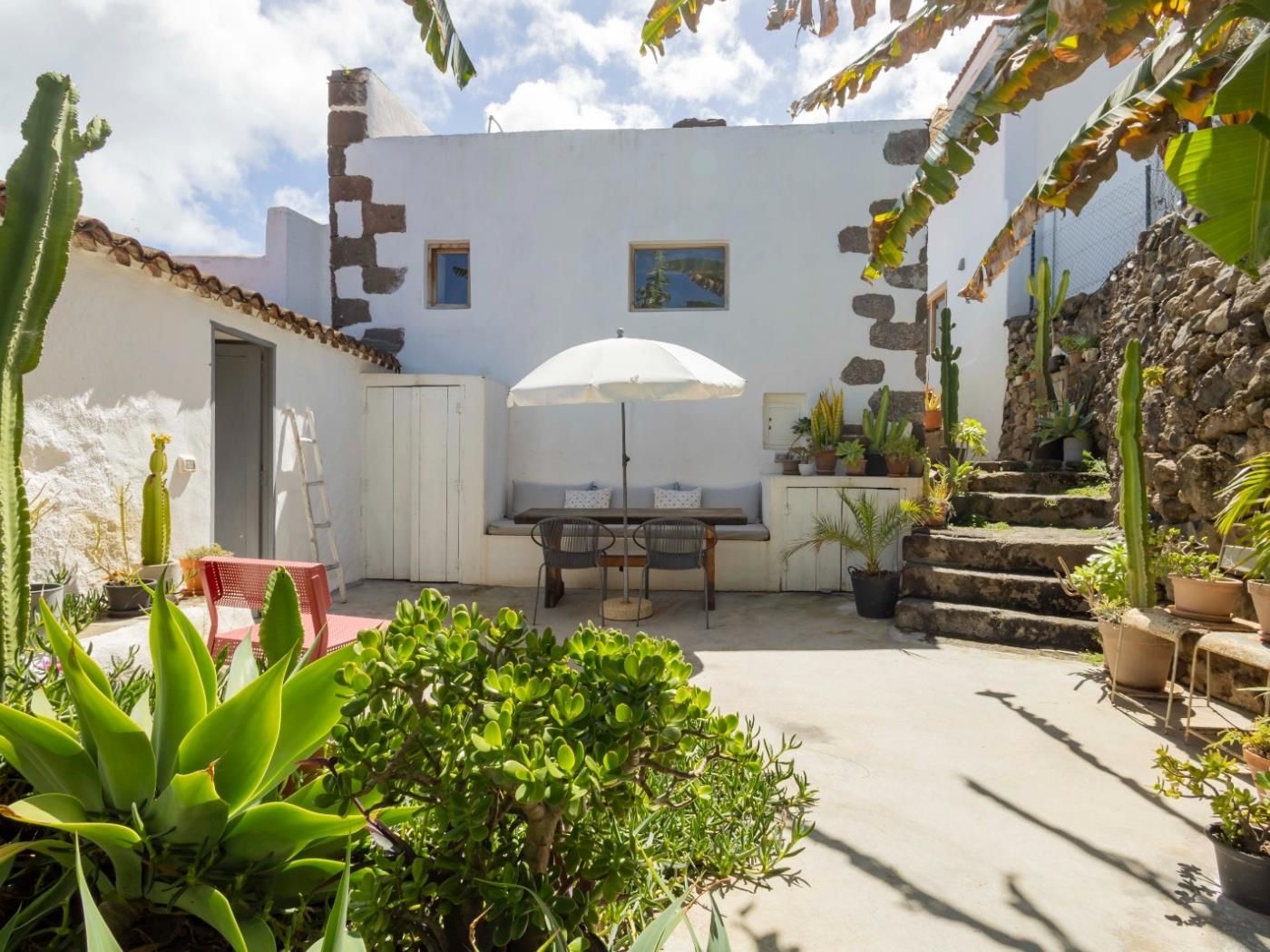 Un patio con una mesa y sillas, rodeado de plantas en macetas y una pared blanca con una ventana pequeña.