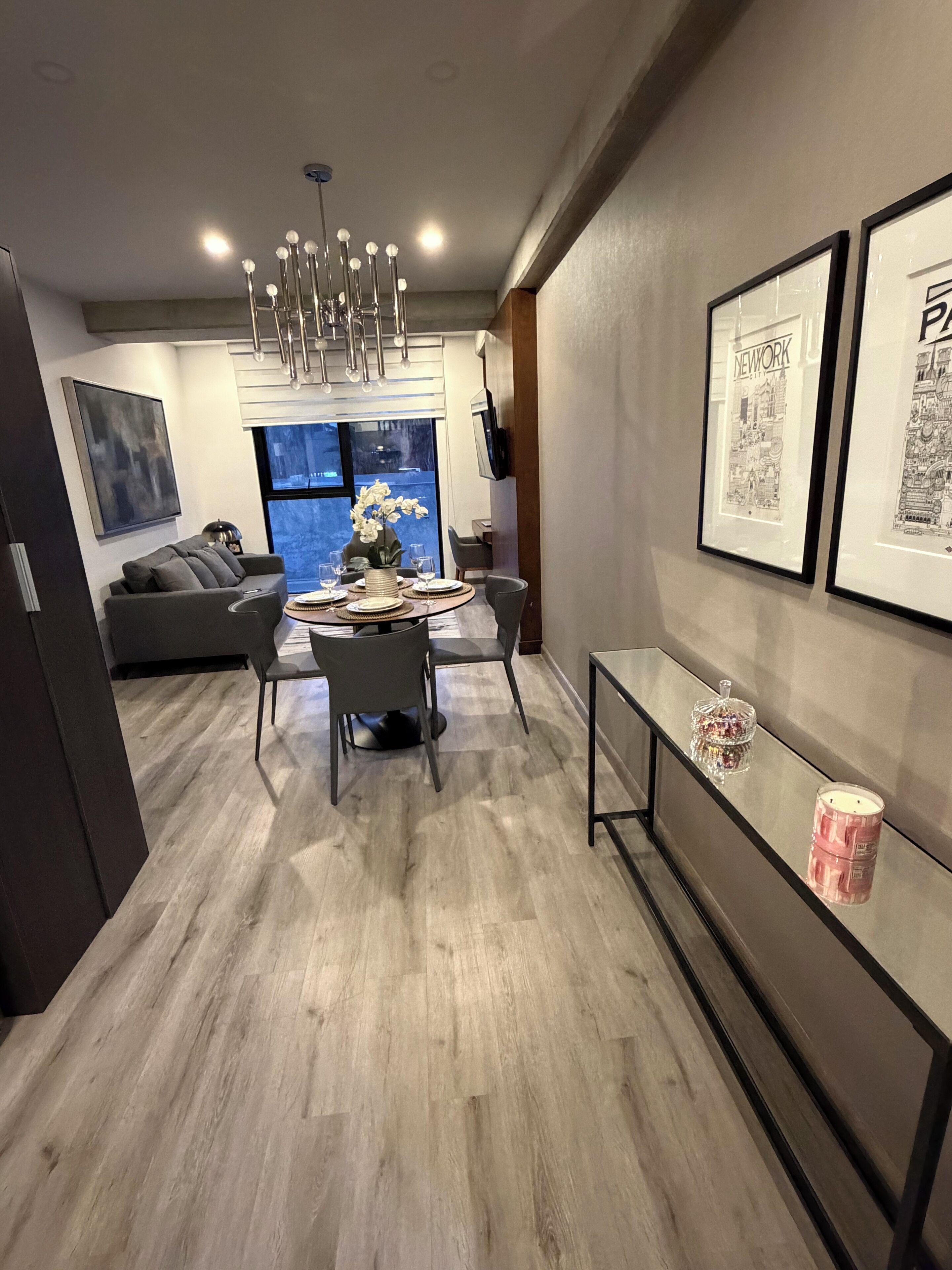 A modern dining area with a round table, chairs, and a chandelier. There is a sofa and a TV mounted on the wall.