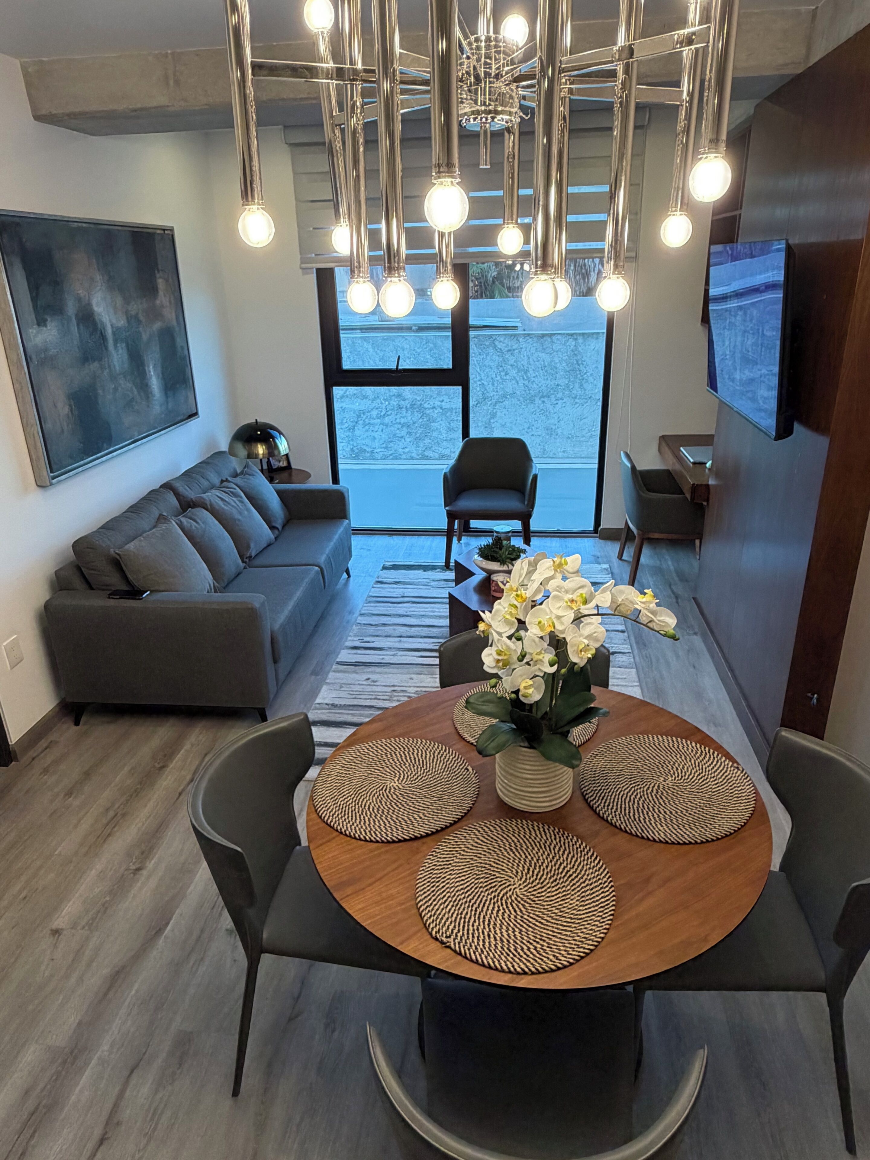 A modern living room with a grey sofa, a dining table with woven placemats, and a chandelier with multiple light bulbs.