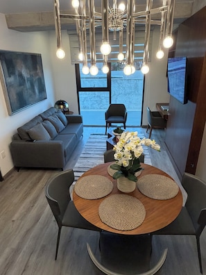 Een moderne woonkamer met een grijze bank, een eettafel met geweven placemats en een kroonluchter met meerdere lichtbollen.