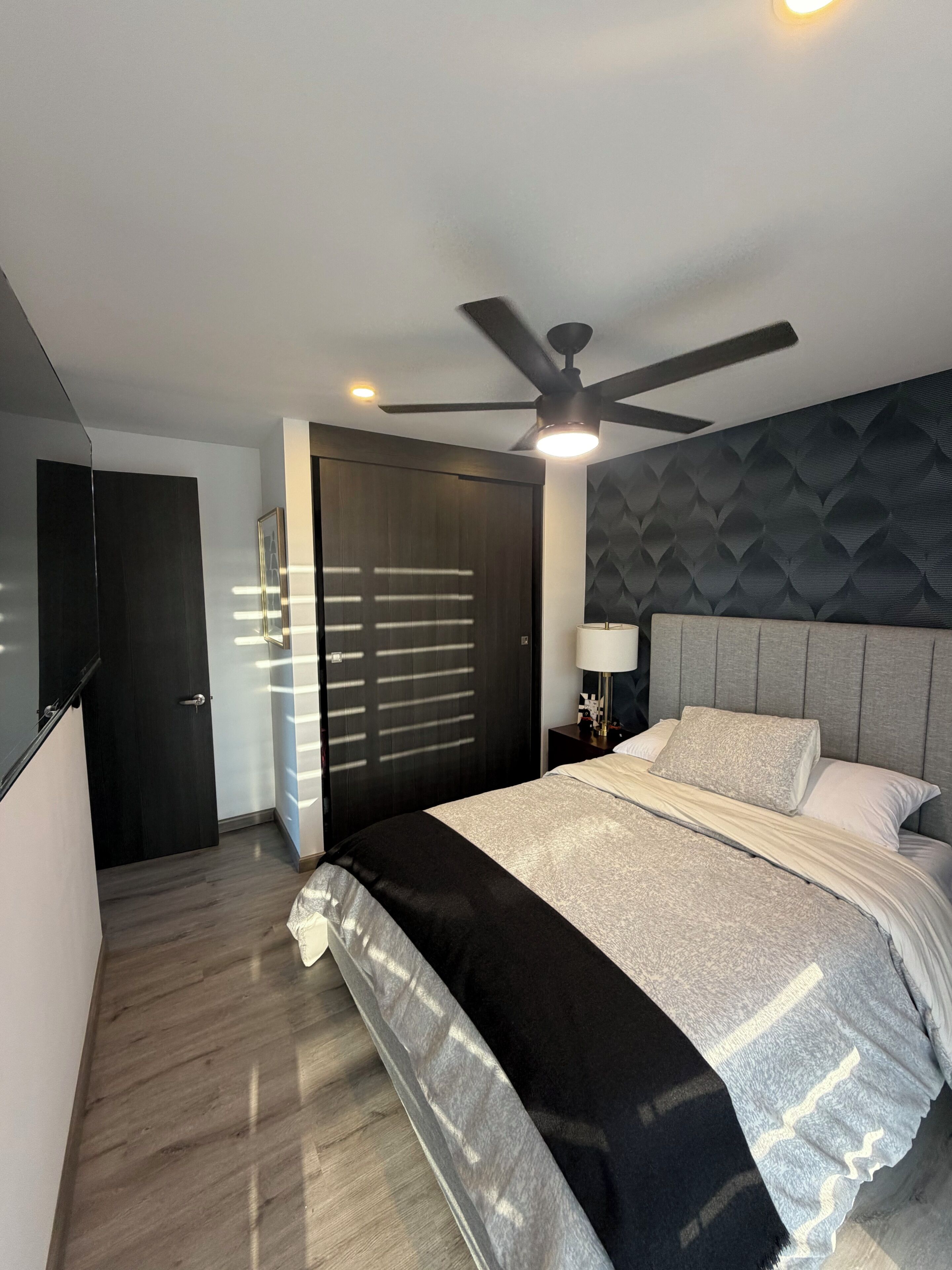 A modern bedroom with a bed, a ceiling fan, a nightstand, and a door.