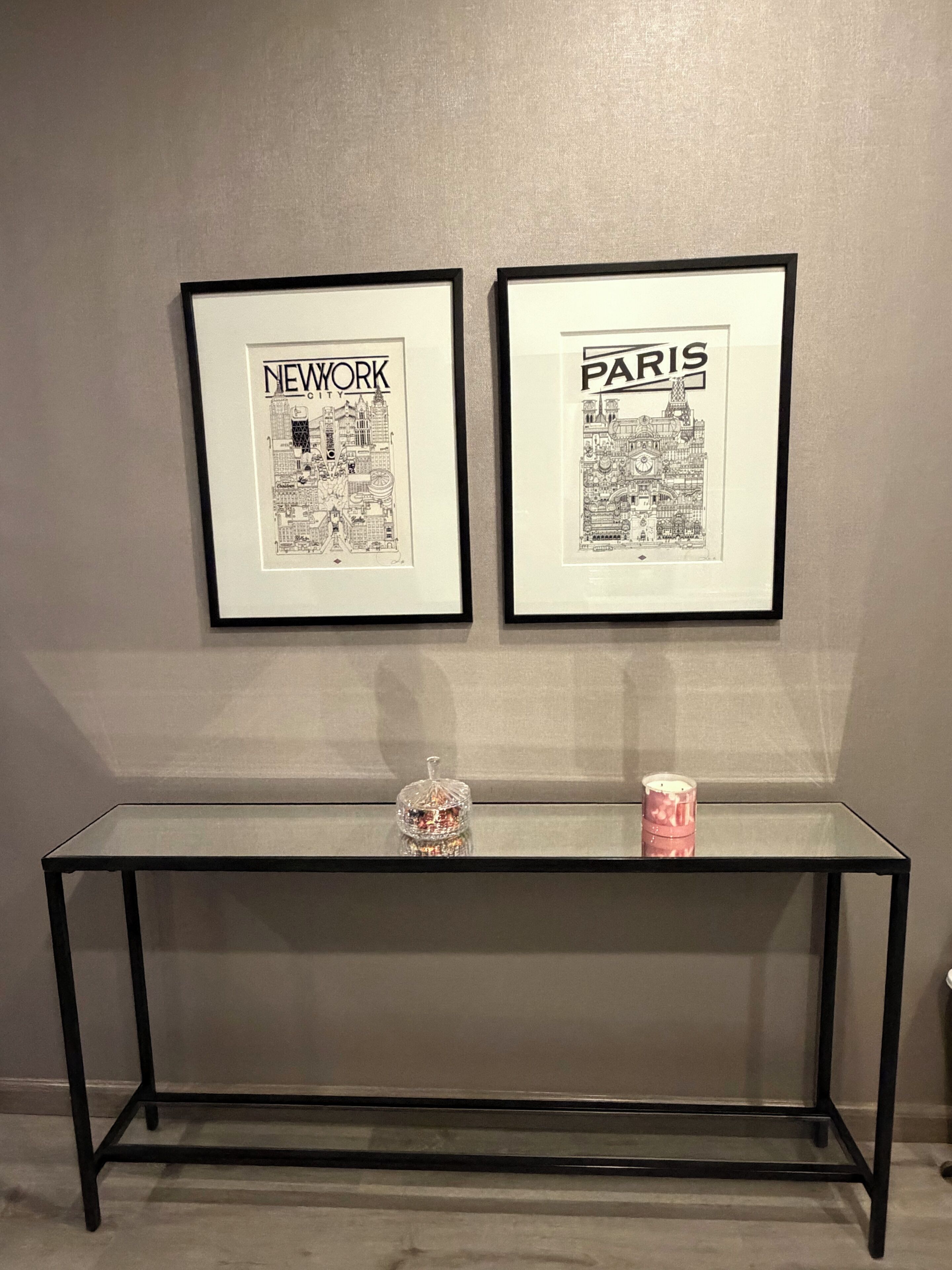 Two framed cityscape illustrations titled 'New York' and 'Paris' hanging on a wall above a glass-top table with a decorative item on it.