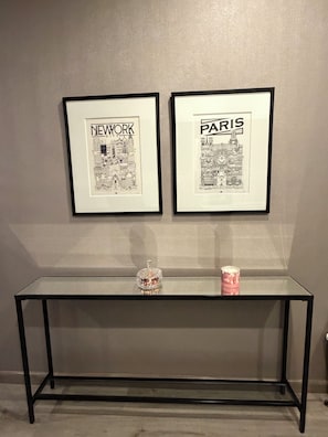 Twee ingelijste illustraties van stadsgezichten, getiteld 'New York' en 'Parijs', hangen aan een muur boven een glazen tafel met een decoratief object erop.
