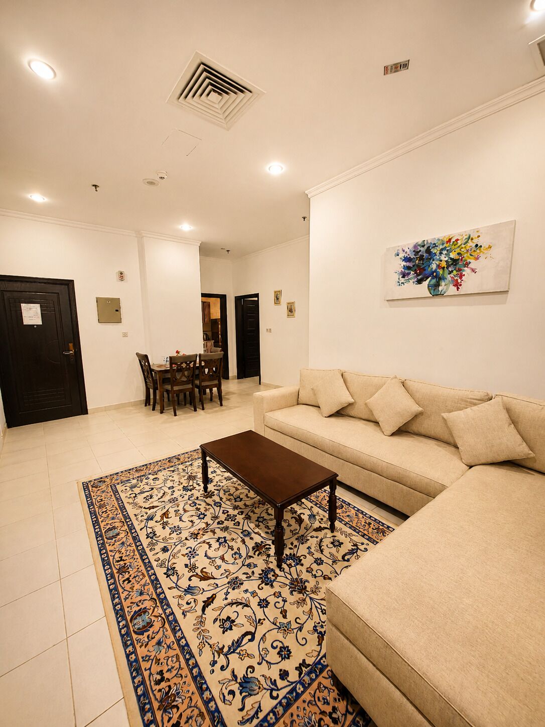 A living room with a beige sofa, a coffee table, and a patterned rug. There is a dining area with chairs and a table in the background.