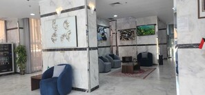 A lobby with marble walls, a large artwork, seating area, and a television screen. - Atlantis Hotel Suite & Apartment (Salmiya)