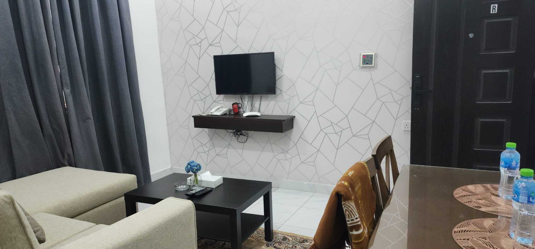 A living room with a sofa, a coffee table, a TV mounted on the wall, a small shelf with phones, and a door labeled 'R'.