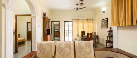 A bedroom with a bed, a ceiling fan, a window with curtains, a dining area with a table and chairs, and a kitchen area.