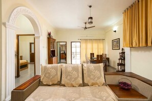 Ein Schlafzimmer mit Bett, Deckenventilator, Fenster mit Vorhängen, Essecke mit Tisch und Stühlen sowie einer Küchenzeile.