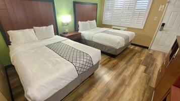 A hotel room with two beds, a nightstand, a wall-mounted lamp, and a window with blinds.