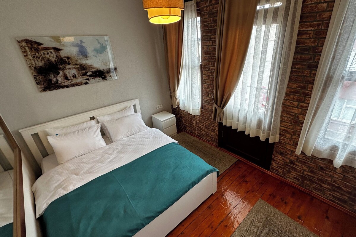 A bedroom with a bed, a nightstand, a wall-mounted lamp, and a window with curtains.