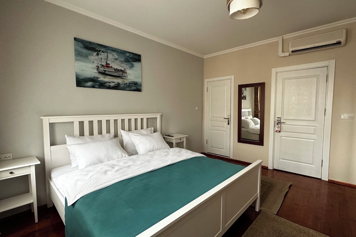 A hotel room with a bed, bedside tables, a painting, a door, and an air conditioning unit.