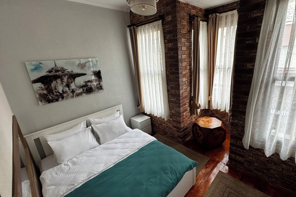 A bedroom with a bed, a nightstand, a wall-mounted painting, and a window with curtains.