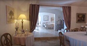 A room with a dining table, chairs, a lamp, and a view into another room with a bed and a chair. - Private Pool Stay in Heart of Luberon (Lagnes)