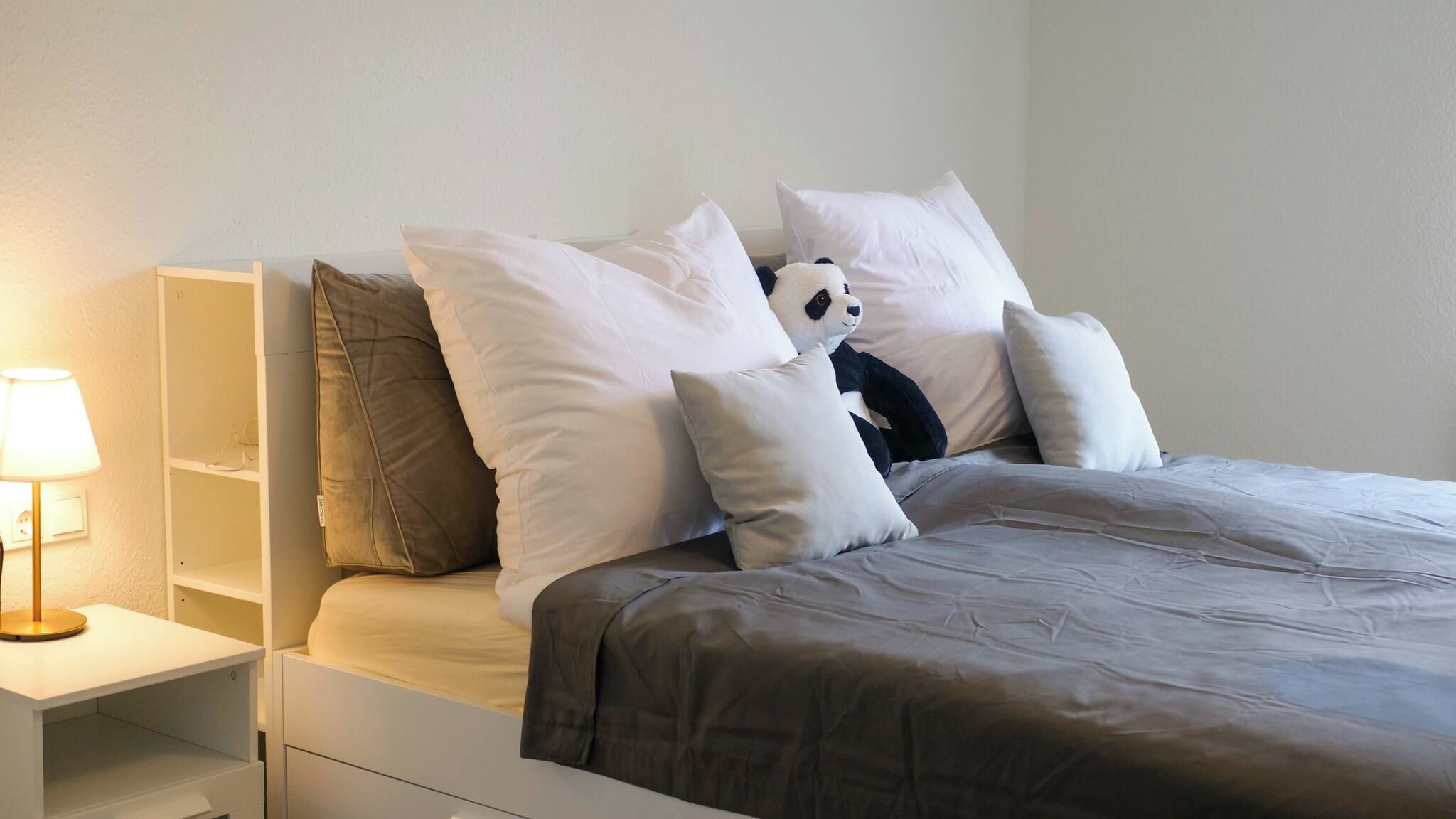 A neatly made bed with a dark brown bedspread and multiple pillows, including a stuffed panda toy.
