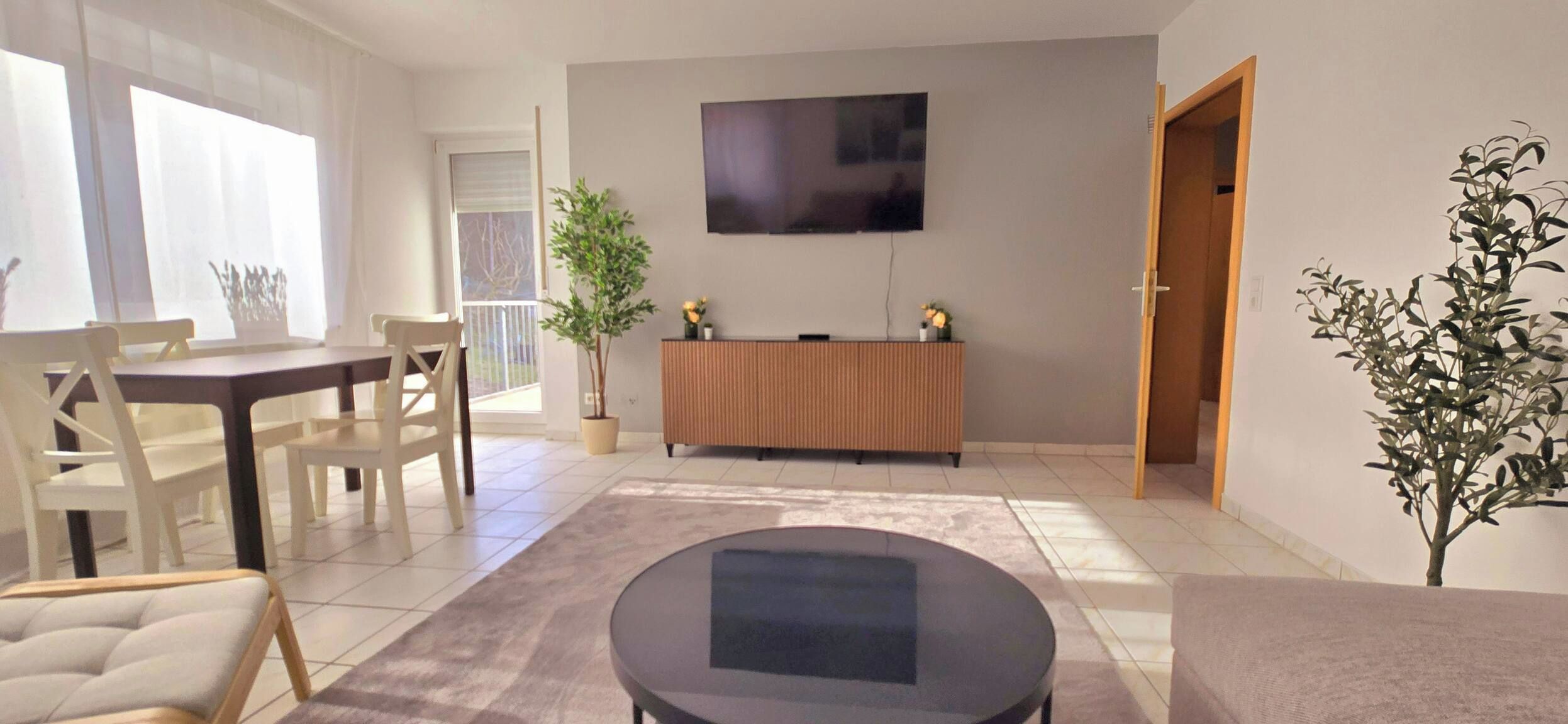 A modern living room with a dining area, a flat-screen TV mounted on the wall, and a potted plant.