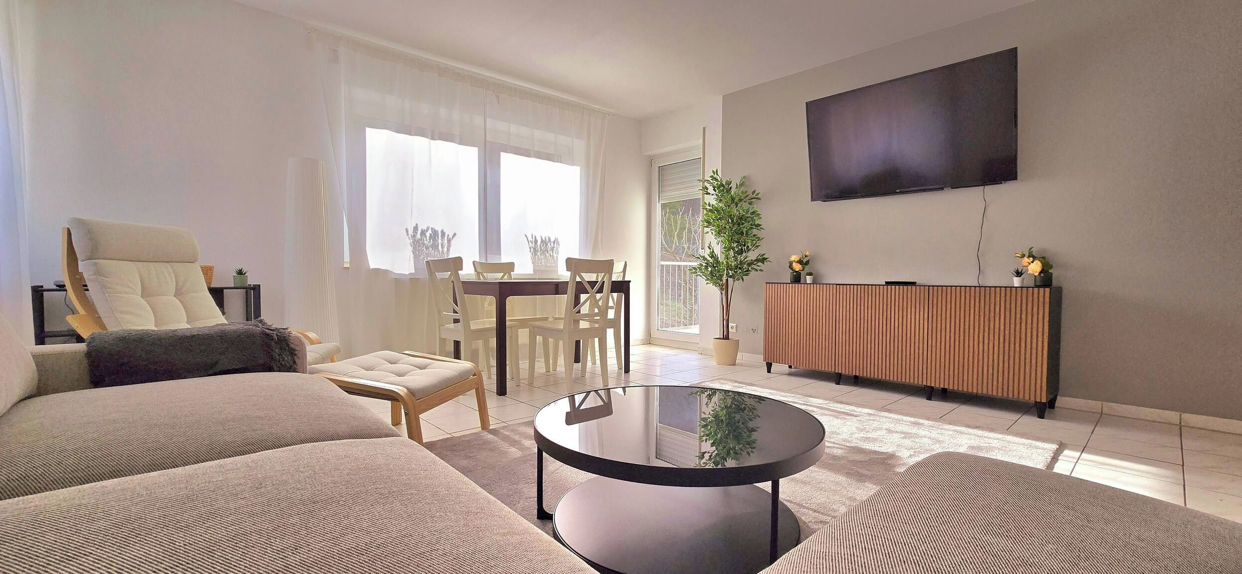 Una sala de estar moderna con un sofá, un sillón, una mesa de comedor, sillas, una mesita de centro, un televisor y una planta en maceta.