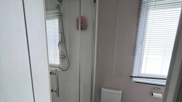 Un baño con cabina de ducha de vidrio, inodoro, radiador y ventana con persianas.
