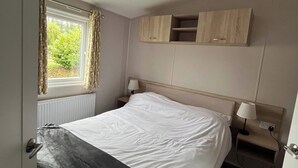 Un dormitorio con cama, ventana con cortinas, mesita de noche con lámpara y un armario de madera sobre la cama.