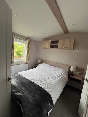 A bedroom with a bed, a window with curtains, a bedside table with a lamp, and a wooden cabinet above the bed. - Clear Water Holidays (Northampton)