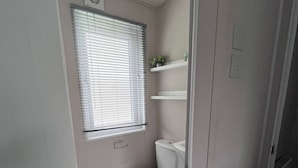 Un baño peque ño con inodoro, una ventana con persianas y un estante con artículos de aseo.