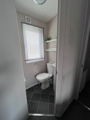 A small bathroom with a toilet, a window with blinds, and a shelf with toiletries. - Clear Water Holidays (Northampton)