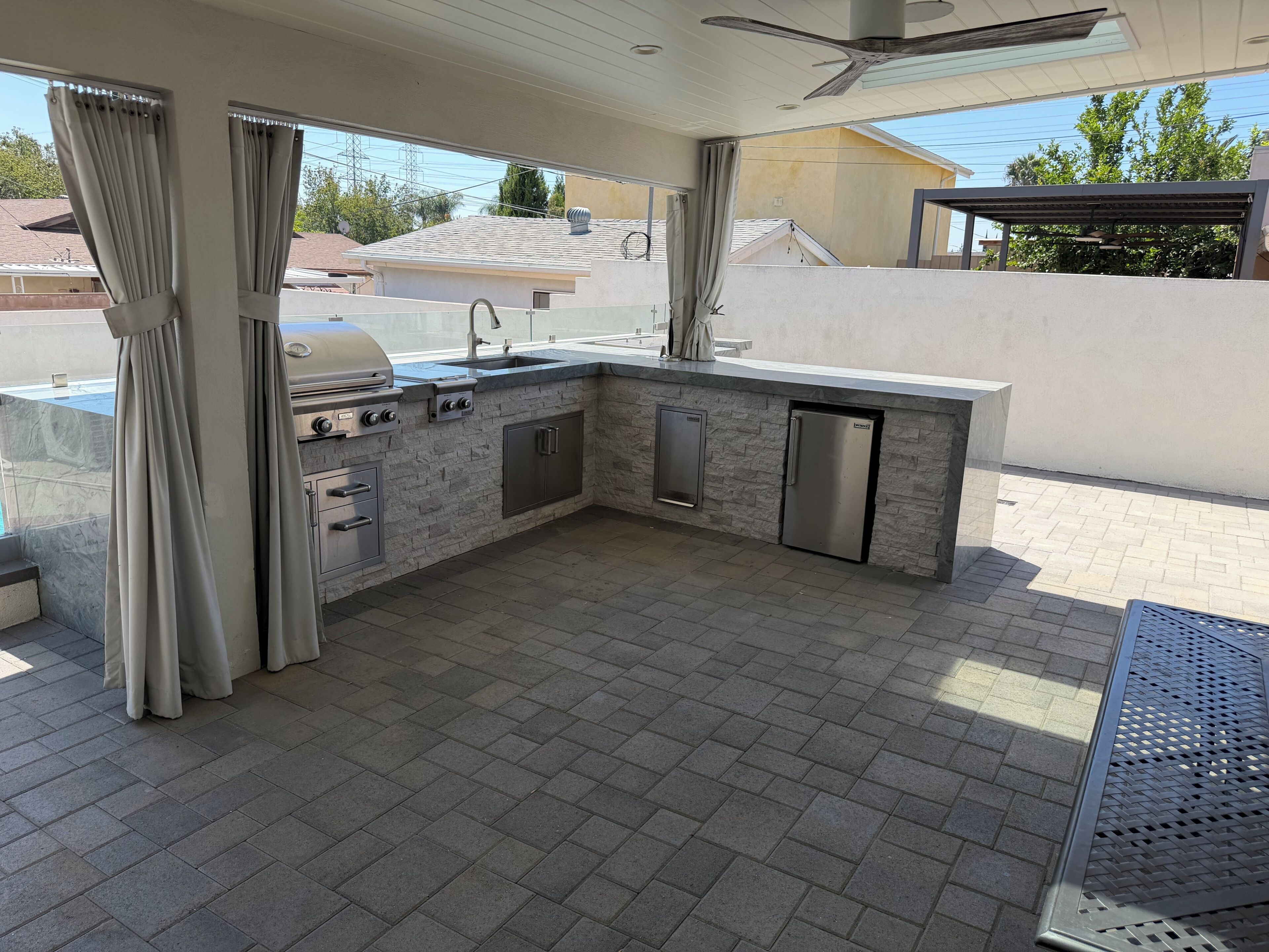 Een buitenkeuken met een barbecue, spoelbak en kastjes, omringd door een patio met een plafondventilator en gordijnen.