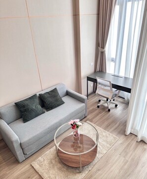 A modern living room with a grey sofa, a round coffee table with a vase of flowers, a desk with a chair, and a large window with curtains. - Duplex 1-bedroom Apartment in Thonglor, Bangkok (Bangkok)