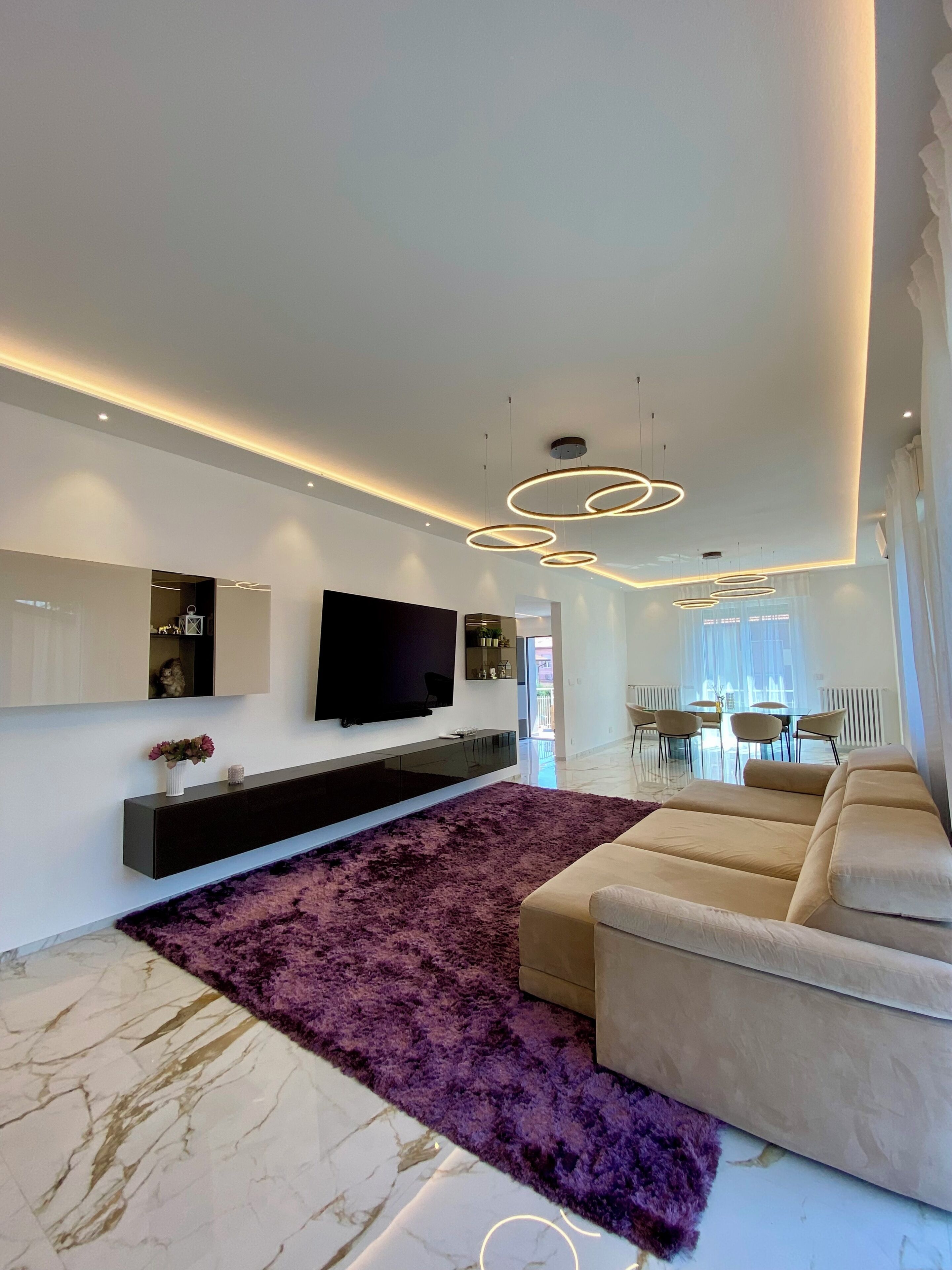 A modern living room with a flat-screen TV, a sofa, a purple rug, and a dining area in the background.