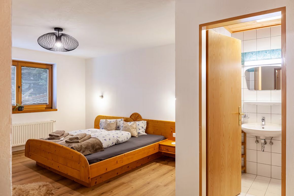 Ein Schlafzimmer mit einem hölzernen Bett, einem Fenster mit Jalousien, einer Deckenleuchte und einem Badezimmer mit Waschbecken und Spiegel.