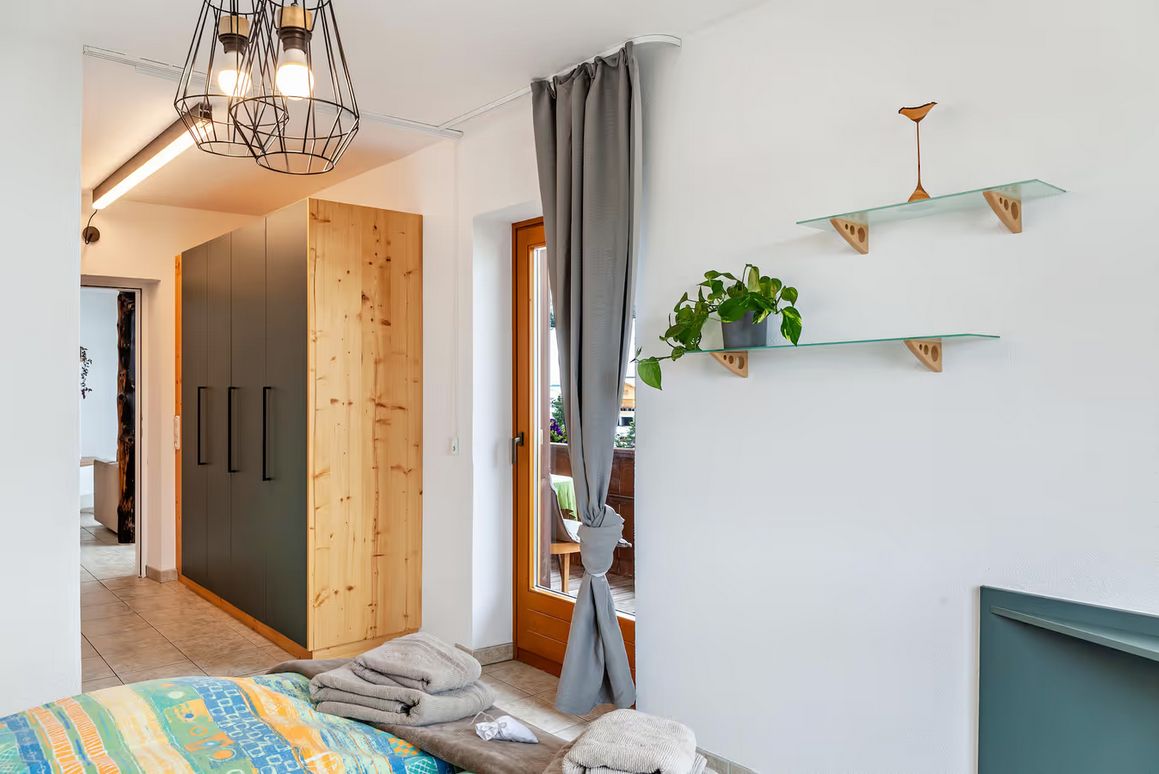 Een slaapkamer met een bed, een kledingkast, een hanglamp, een raam met gordijnen en een wandplank met een plant.