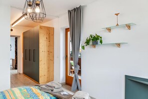 A bedroom with a bed, a wardrobe, a hanging light fixture, a window with curtains, and a wall-mounted shelf with a plant.