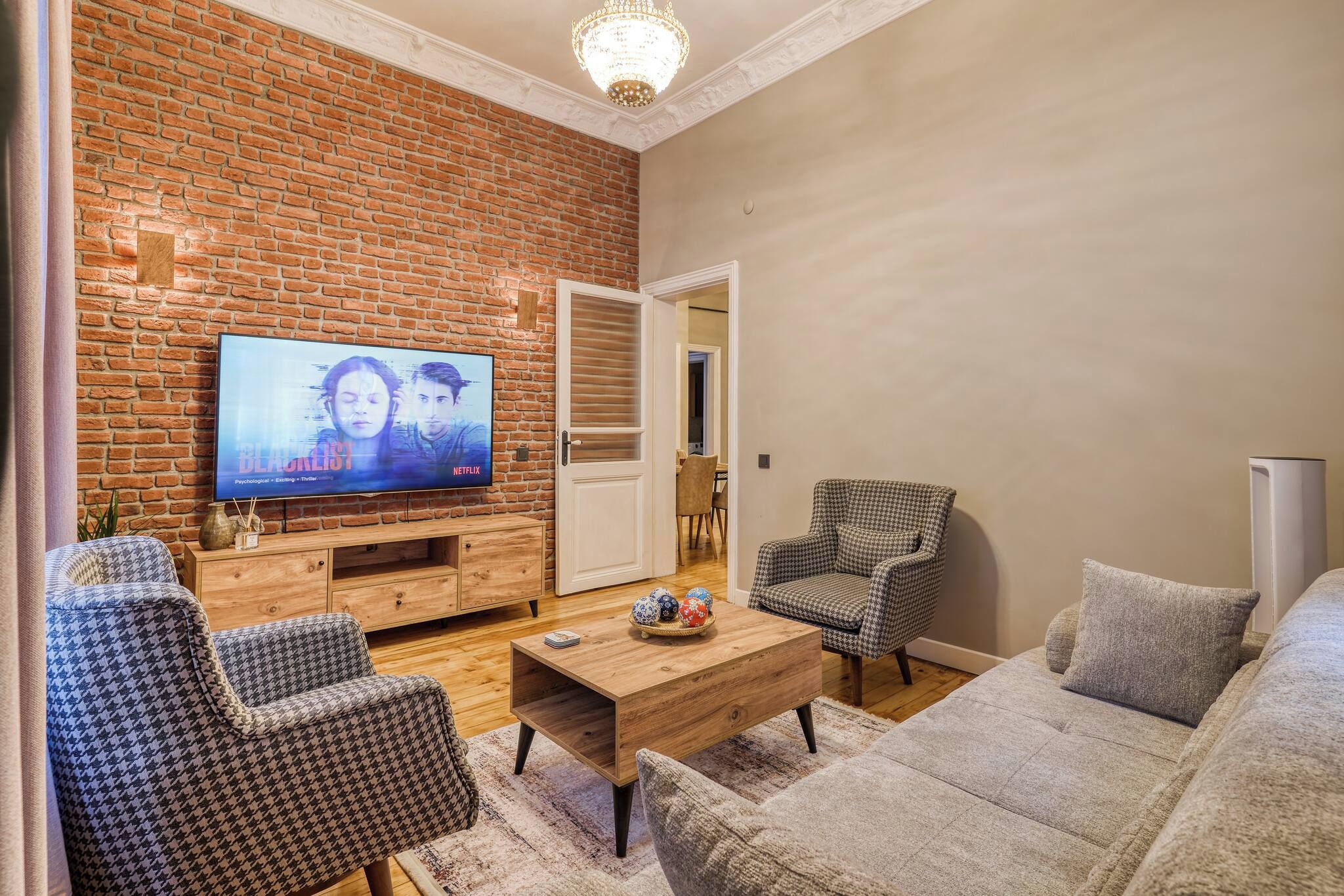 A living room with a brick wall, a television, a sofa, two patterned armchairs, a coffee table, and a door leading to another room.