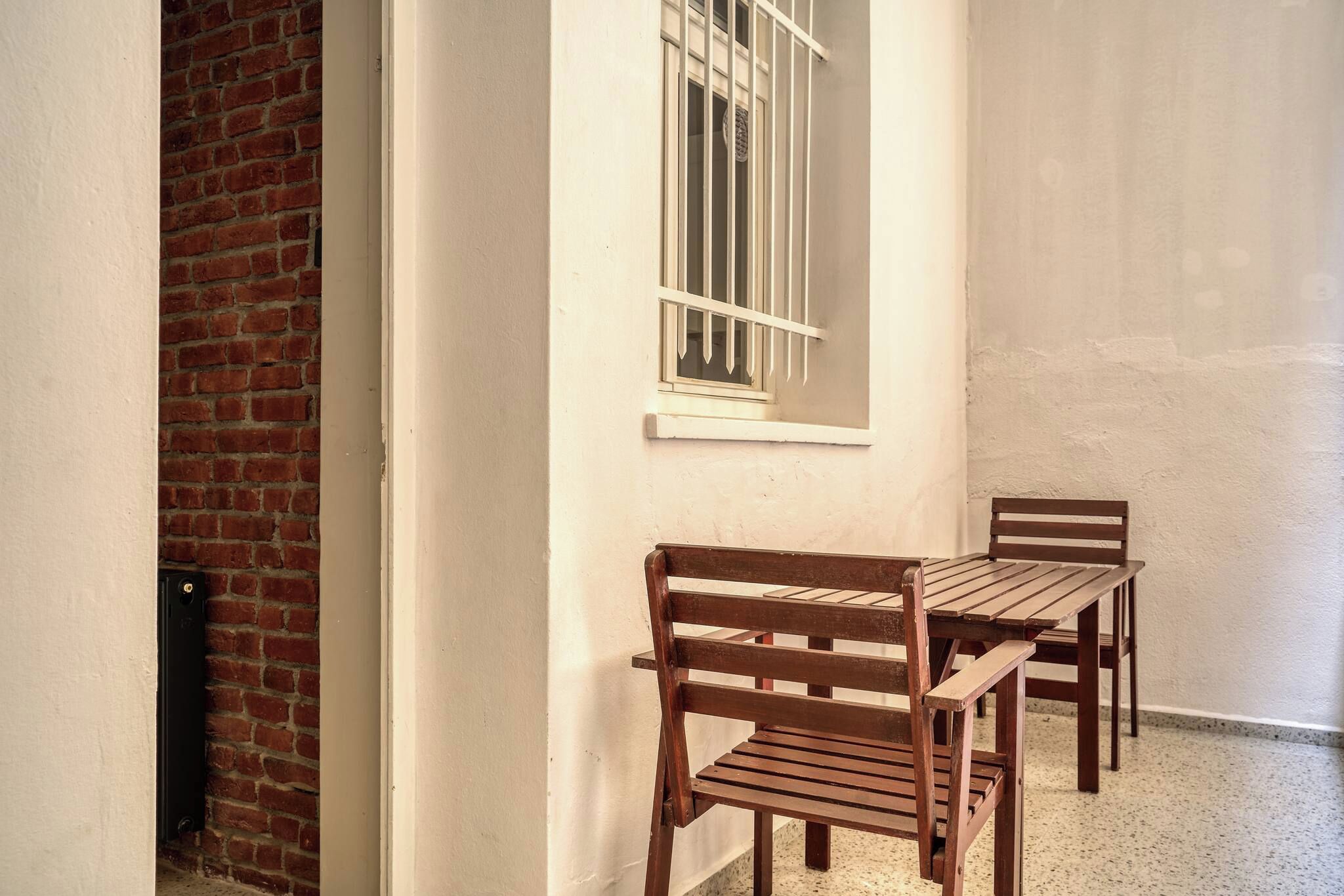 A narrow corridor with a brick wall on one side, a white wall with a window on the other, and a wooden bench and table set against the wall.