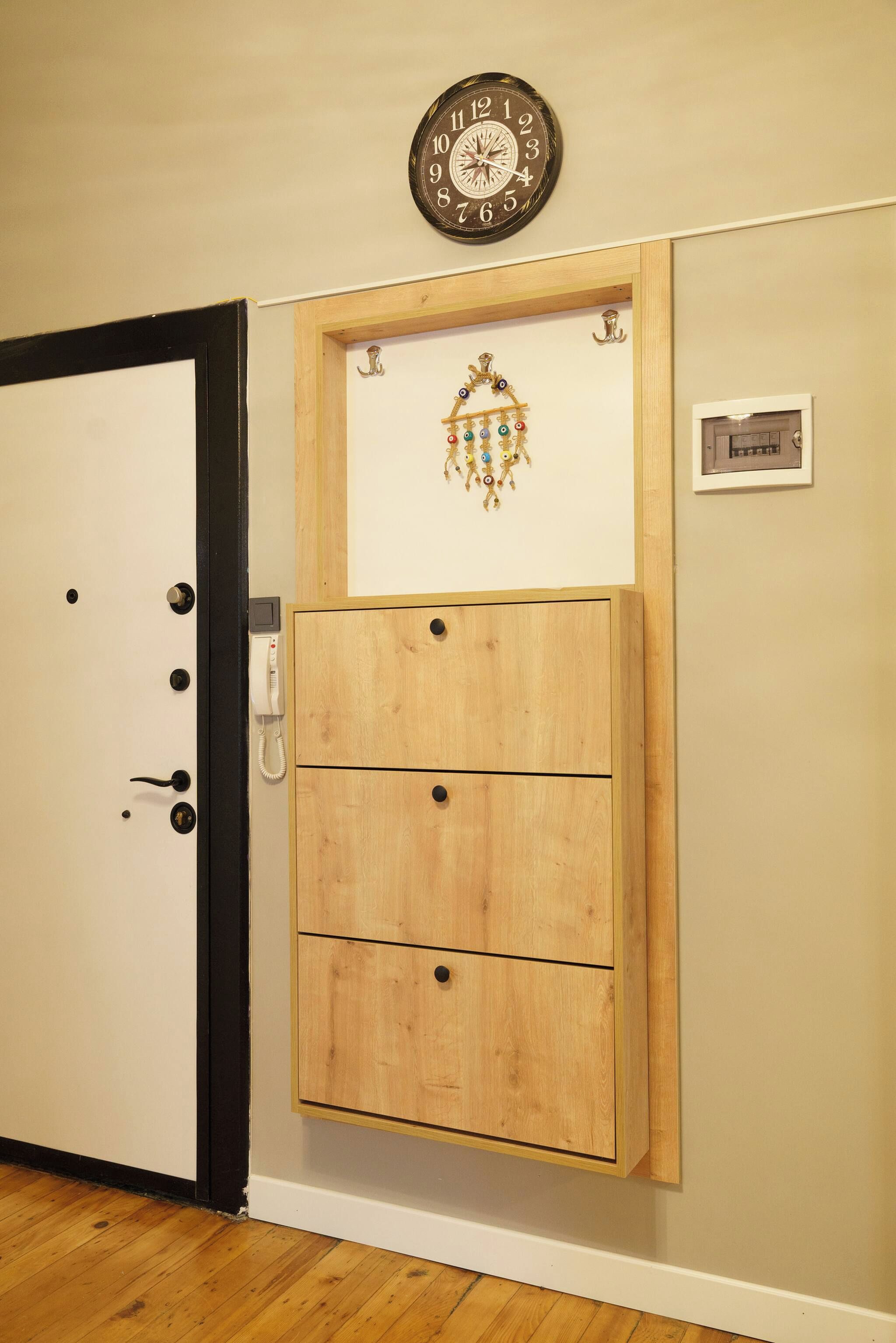 A wooden cabinet with three drawers, a wall clock, and a small plaque on a beige wall.