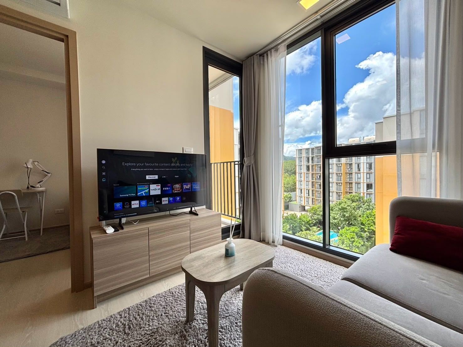 Et moderne hotelværelse med et stort fladskærms-tv, der viser en streamingtjenestes brugerflade, en sofa, et lille bord og udsigt over bybilledet gennem store vinduer.