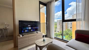 Et moderne hotelværelse med et stort fladskærms-tv, der viser en streamingtjenestes brugerflade, en sofa, et lille bord og udsigt over bybilledet gennem store vinduer.