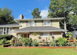 A two-story house with a front porch, a brick chimney, and a garden with various plants and flowers.