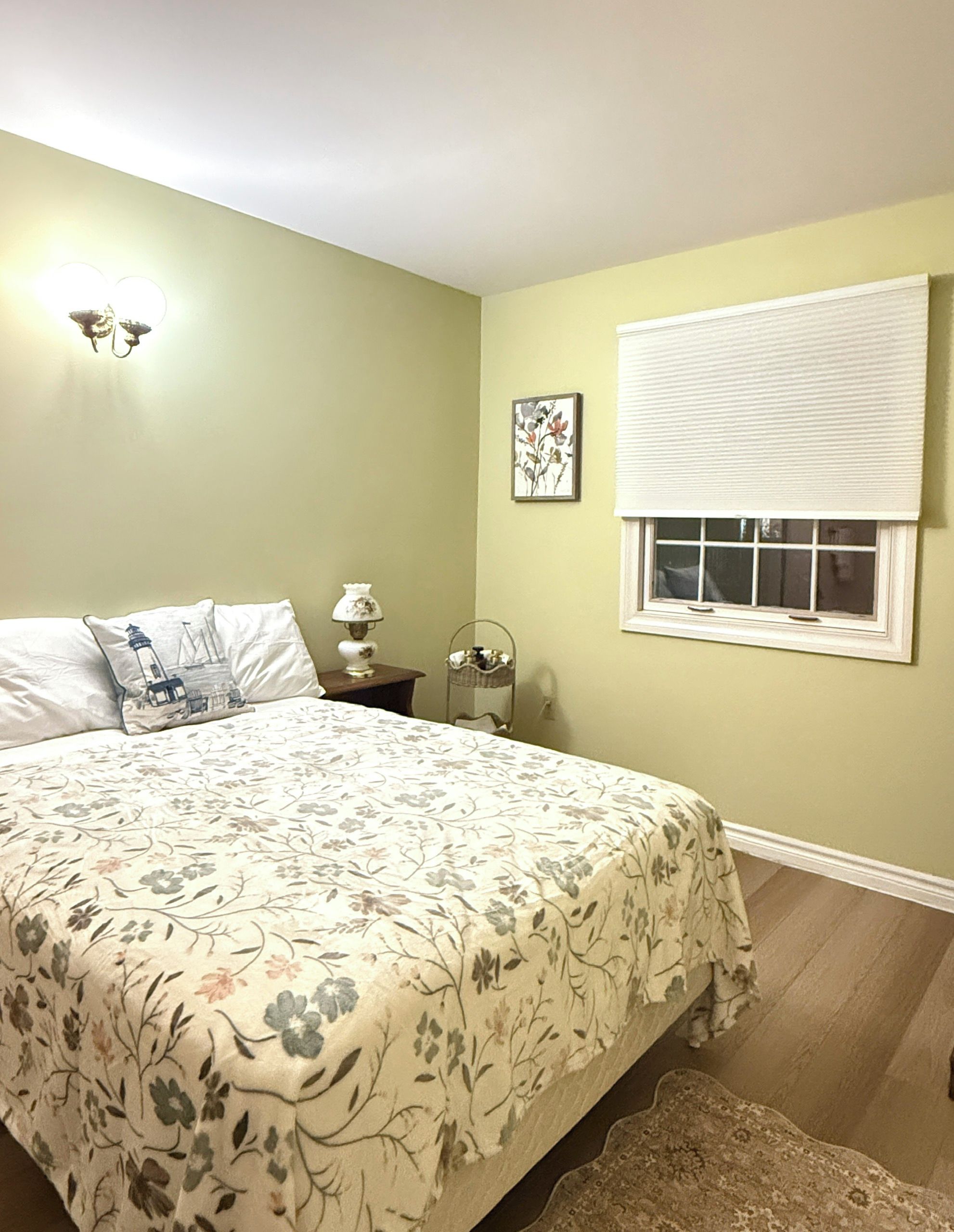 A bedroom with a bed, a nightstand, a lamp, a window with blinds, and a wall-mounted light fixture.
