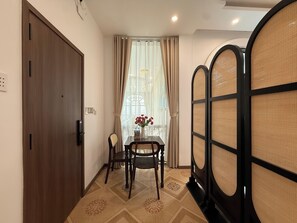 A room with a wooden door, a table with chairs, a window with curtains, and a decorative wall panel. - Maison - Product by Qunh Khang (Tân Sơn Hòa)